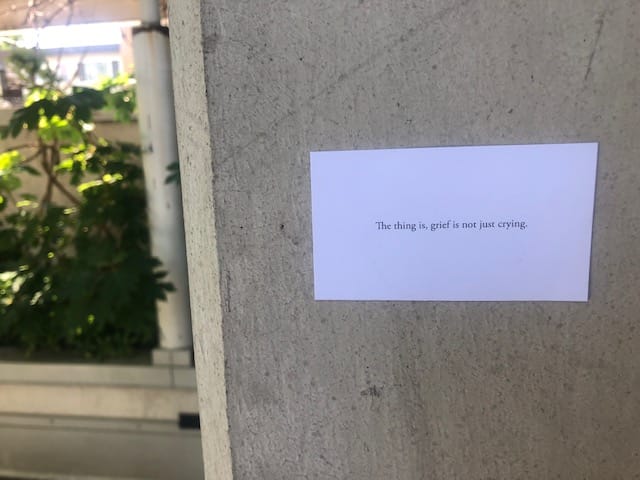 A piece of paper is on a cement wall, in the background there is some greenery. The paper reads: The thing is, grief is not just crying. Photo by Adrienne Wong.